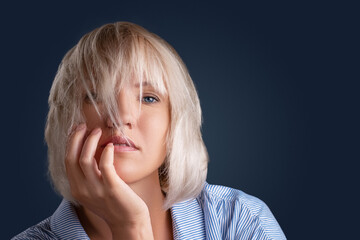 Young stylish caucasian girl on a dark background with beautiful blue eyes, plump lips looks at the camera. Blond hair covers part of the face. Hand and fingers touch her face.