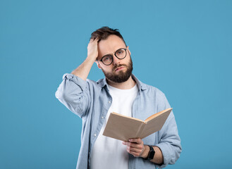 Studying is hard. Young bearded guy having problem understanding textbook on blue studio background