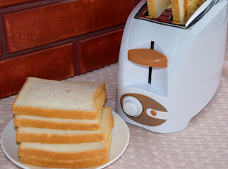 There is a toaster in the kitchen against a red brick wall. Fried toast bread emerges from the toaster. Next to the toaster is a plate of toast bread for toasting.