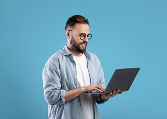 Confident young guy in glasses using laptop pc for online work or communication on blue studio...