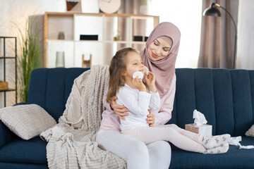 Lovely caring Muslim mom, sitting on blue sofa at home and holding her little daughter on knees....