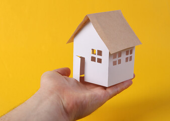 Fototapeta na wymiar Hand holds mini paper home figurine on yellow background. Protection concept, security