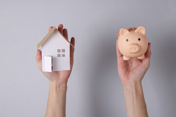 Home buying concept, save money. Hand holds mini house and piggy bank on gray background