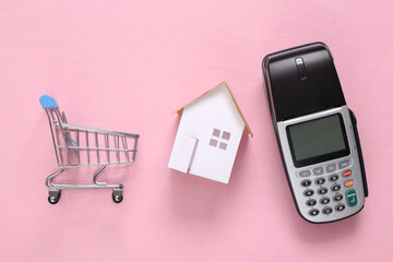 Home buying concept. Mini house, payment terminal and shopping trolley on pink blue background. Top view