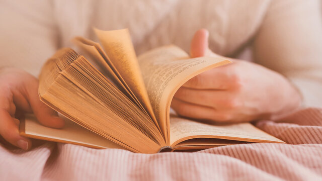 A man in a white sweater is lying on a bed with a soft pink blanket and flipping through the pages of an old interesting book. Rest at home. Self-development.