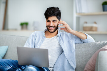 Cheerful arab man using laptop and talking on phone at home, sitting on sofa in living room