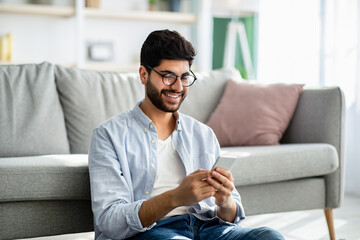 Arab guy texting and browsing internet on smartphone, resting at home, leaning on sofa