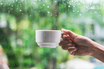 Hand holding cup of coffee with raindrop and defocused green nature background, Rainy season concept
