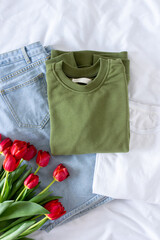 Female trendy minimalism clothes flat lay on white sheet background, women set of blue jeans, white t shirt and khaki green sweatshirt with red tulips top view, fashion blogger content