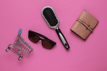 Shopping concept. Fashionable women's accessories. Leather wallet, comb, sunglasses, mini shopping trolley on pink background. Top view