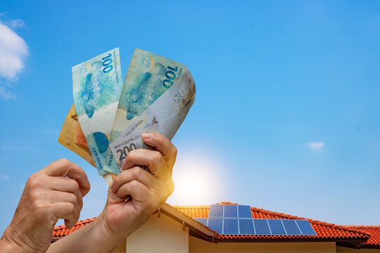 Solar Energy Economy, holding Brazilian money in front of the photovoltaic panel on a roof, sunset background.