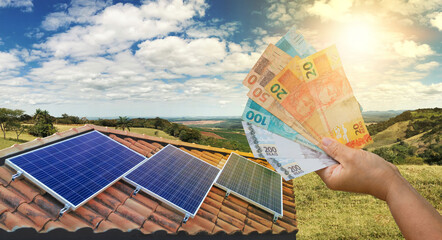 Solar Energy Economy, holding Brazilian money in front of the photovoltaic panel on a roof, sunset background.