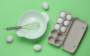 Obraz na płótnie Canvas Cooking concept. Empty white bowl with whisk, egg tray on green studio background. Top view.
