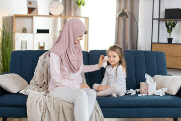 Front view of young pretty mother, Muslim woman in hijab, sitting on blue couch at home and wiping face of her cute little 3 years old daughter with paper napkin. Morning hygiene routine at home