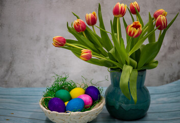 Turquoise ceramic vase with yellow-red tulips and colorful Easter eggs in a bowl, wooden tray