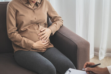 Young asian pregnant woman holding her belly while gynecologist notes the symptoms that the pregnancy is explaining about the unborn child.