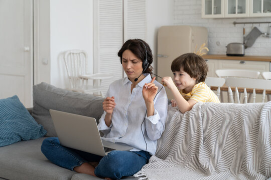 Nervous freelancer mother sitting on couch at home office during lockdown, working on laptop. Little child distracts from work, taking off headphones, making noise and asking attention from busy mom
