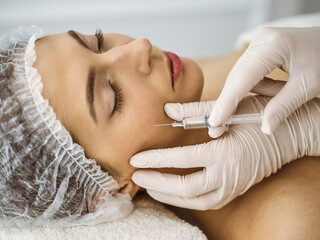 Naklejka premium Beautician doing beauty procedure with syringe to face of young brunette woman in sunny clinic. Cosmetic medicine and surgery, beauty injections
