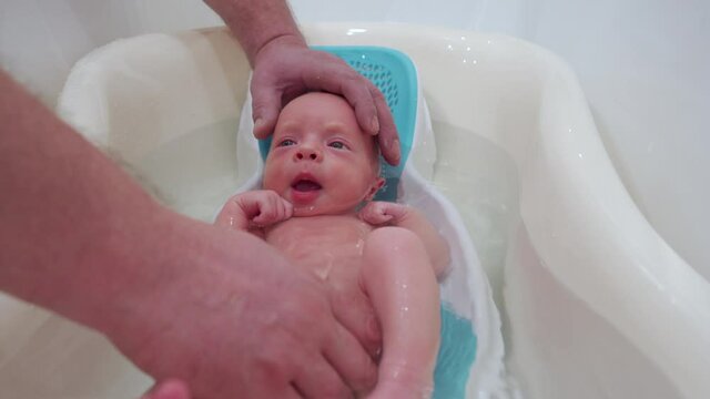 Bathing newborn baby in a baby bath, father bathing his 3-week-old son, infant in baby bath seat support.