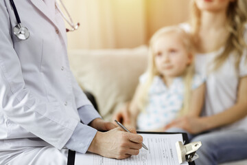 Fototapeta premium Doctor and patient. Pediatrician using clipboard while examining little girl with her mother at home. Sick and unhappy child at medical exam