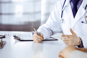 Unknown male doctor and patient woman discussing something while sitting in clinic and using clipboard. Best medical service in hospital, medicine, pandemic stop