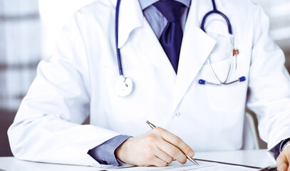 Unknown male doctor sitting and working with clipboard in clinic at his working place, close-up. Young physician at work. Perfect medical service, medicine concept