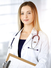 Young optimistic woman-doctor is holding a clipboard in her hands, while standing in a sunny clinic. Portrait of friendly female physician with a stethoscope. Perfect medical service in a hospital