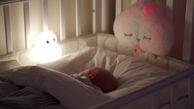 newborn baby boy sleeping in bed in the evening, 3-week-old infant in light of a night lamp, night light with lullaby light show.