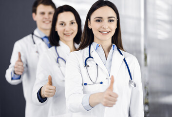 Fototapeta premium Group of young cheerful doctors is standing as a team with thumbs up in a hospital office and is ready to help patients. Medical help, insurance in health care, best disease treatment and medicine