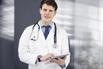 Young friendly doctor is checking some information on his computer tablet. Portrait of a professional physician at work in a clinic. Medicine concept