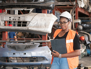 African woman warehouse automotive parts worker wears a safety helmet takes note on the clipboard among the old engines automotive spare parts. .