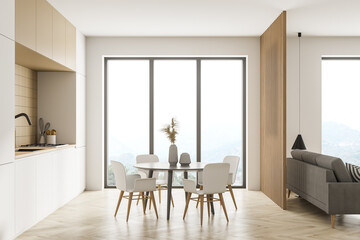 Modern kitchen interior, dining area and living room space with panoramic windows with countryside and hills view. Furnished by table and chairs for eating, beige sofa.