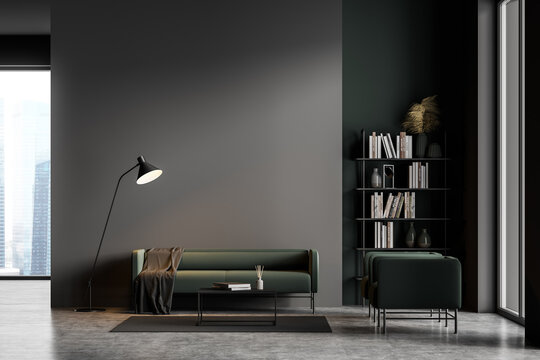 Dark grey modern living room interior with panoramic windows furnished by green sofa and armchairs, coffee table, bookshelf, lamp. Concrete floor.