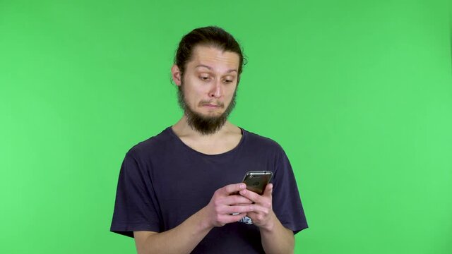The man reads something on his phone, which shocks him, and writes back a text message on his phone. A darkhaired man with a beard in a black Tshirt in the studio on a green screen. Close up.