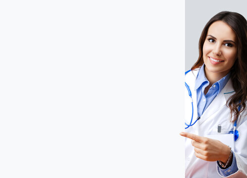 Portrait image of happy smiling female doctor pointing blank signboard with copy space empty area for some text, over grey. Young brunette beautiful woman in medical, clinic, healthcare studio concept