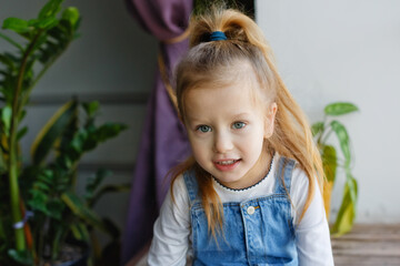 A little blonde girl with blue eyes in a denim jumpsuit is sitting on a large wooden window sill near the window.