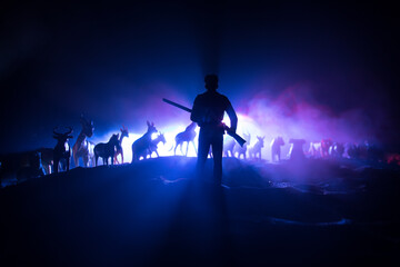 Silhouette of a man (hunter) with rifle standing against group of animals in colorful dark backlight. Decorated with miniatures. Selective focus