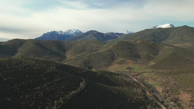 Aerial view over a railway track and road passing through a valley in the Balagne region of Corsica with the snow capped peaks of the Asco mountains and Monte Padru in the distance