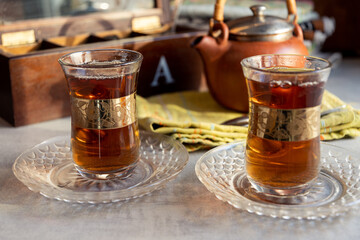 Tea in glass Turkish cups with a gold pattern. A ceramic teapot on a kitchen napkin and a wooden tea box.