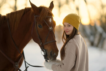 Side portrait of young woman and brown horse. Woman with long hear in yellow cap holding snaffle of horse