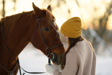 Winter side portrait of young woman and brown horse. Woman with long hear in yellow cap holding snaffle of horse