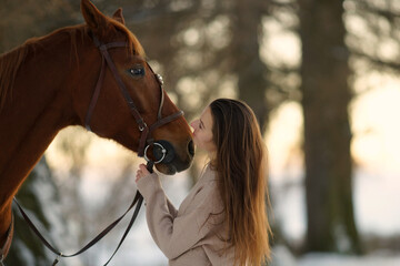 Side portrait of young woman and brown horse at winter sunset. Woman with long hear in sweater standing near big horse