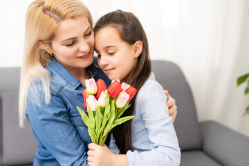 Child daughter is congratulating mom and giving her flowers tulips. Mum and girl smiling and hugging. Family holiday and togetherness.