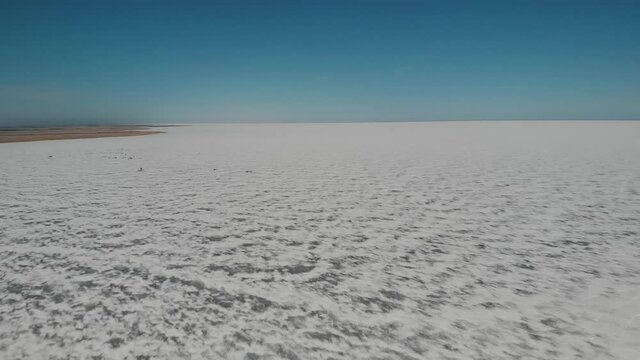 Drone flying slowly ahead with a view of a vast frozen lake covered with snow drifts under a bright blue sky
