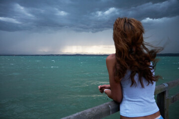Young woman stands on a parapet on a pier and looks over the water. In the distance it is raining. She wears a short skirt and has long hair blowing in the wind. The focus is on the background. 
