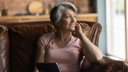 Nostalgia. Peaceful smiling elderly aged female grandma sit on comfy couch hold tablet dream distracted from gadget screen. Pensive older hispanic woman rest of pad computer lost in positive thoughts