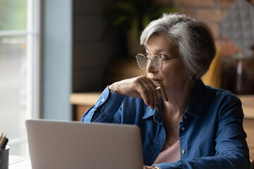 Old age and tech. Thoughtful aged latin female sit at desk work online by laptop from home office....