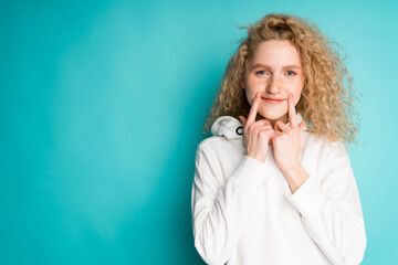 Cute young girl with curly hair with index fingers touches her mouth to make a smile