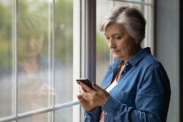 Important message. Serious aged latin female pensioner stand by picture window at home hold cell text in messenger app. Focused older woman thinking on email received from bank write answer. Copyspace