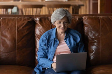 Aged user. Confident elderly latin lady wearing glasses sit on sofa hold computer on knees type...
